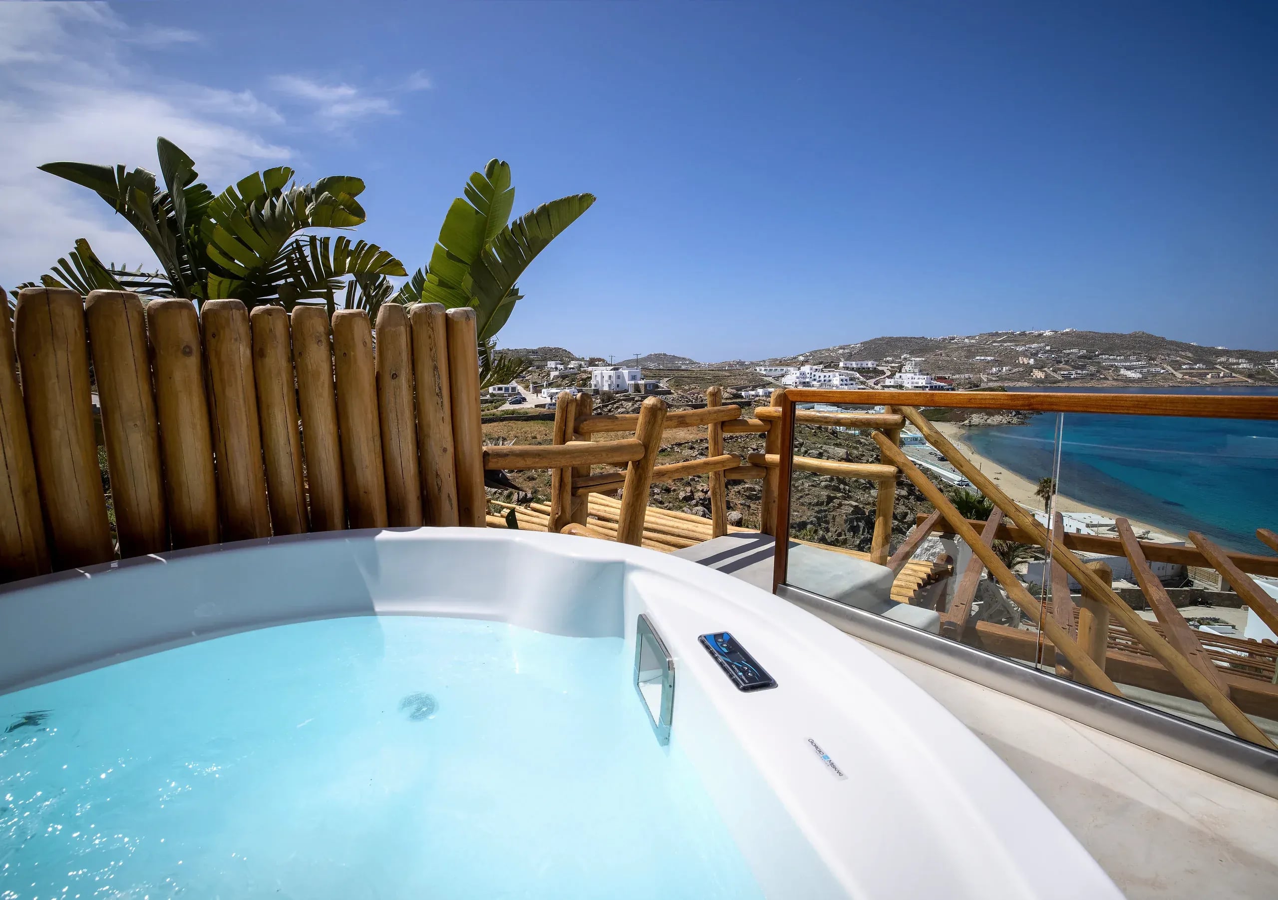 An outdoor hot tub behind a timber pole screen, with planting and the bay beyond